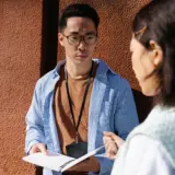 Doi tineri cu trăsături asiatice, un bărbat și o femeie, discută în aer liber lângă un perete texturat de culoare maro-roșcat