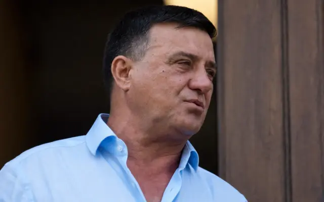 A close-up of Niculae Bădălău in a blue shirt standing in front of a wooden door with signs