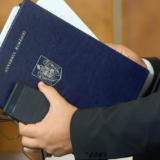 Un bărbat la costum ține sub braț o mapă de culoare bleumarin pe care scrie Guvernul României și are stema țării