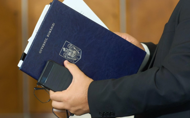 Un bărbat la costum ține sub braț o mapă de culoare bleumarin pe care scrie Guvernul României și are stema țării