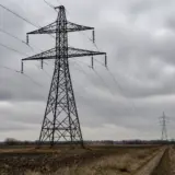 Doi stâlpi de înaltă tensiune din metal, unul în prim-plan și unul în depărtare, conectați prin cabluri electrice care traversează un câmp arat