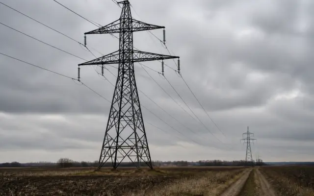 Doi stâlpi de înaltă tensiune din metal, unul în prim-plan și unul în depărtare, conectați prin cabluri electrice care traversează un câmp arat