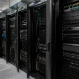 O perspectivă dintr-un centru de date prezentând mai multe rack-uri negre parțial deschise, pline cu servere, switch-uri și cabluri de rețea