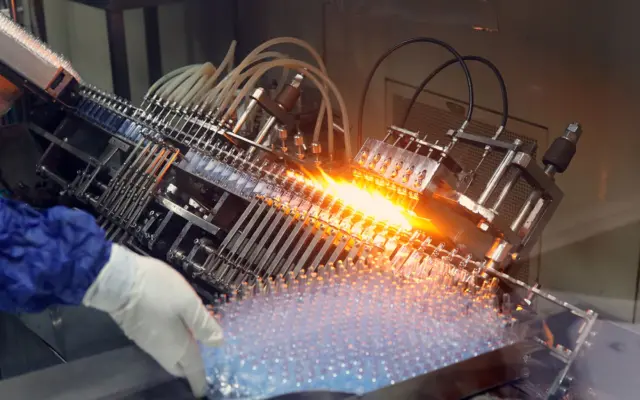 O mașină industrială complexă într-un proces de fabricație, cu flăcări care sigilează flacoane de sticlă pe o bandă rulantă