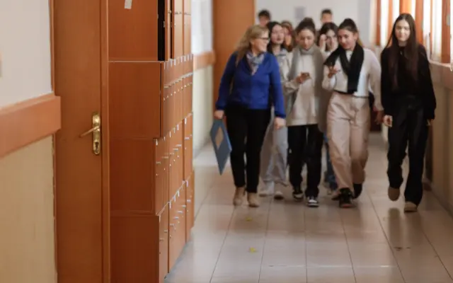 Un grup de elevi și un profesor merg pe un coridor de școală, pe lângă un rând de dulapuri metalice de culoare portocalie