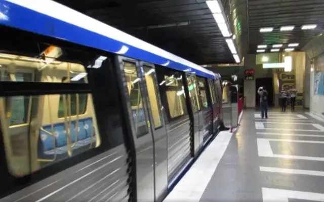 Un tren de metrou model CAF oprit în stația de metrou Piața Romană din București, cu câțiva călători pe peron și luminile stației aprinse