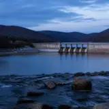 O imagine de ansamblu cu un baraj de beton situat între dealuri împădurite, cu un lac de acumulare în spatele său și un curs de apă cu bolovani în prim-plan