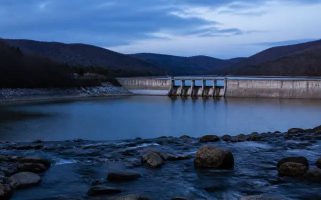 O imagine de ansamblu cu un baraj de beton situat între dealuri împădurite, cu un lac de acumulare în spatele său și un curs de apă cu bolovani în prim-plan