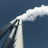O perspectivă de jos în sus către trei hornuri industriale înalte, de culoare gri, din care iese abur sau fum alb dens pe un cer albastru curat