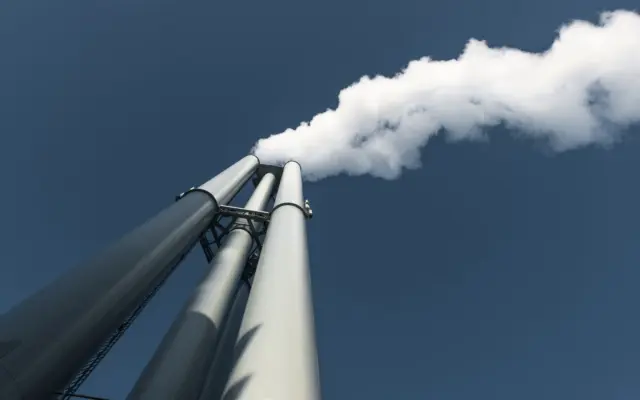 O perspectivă de jos în sus către trei hornuri industriale înalte, de culoare gri, din care iese abur sau fum alb dens pe un cer albastru curat