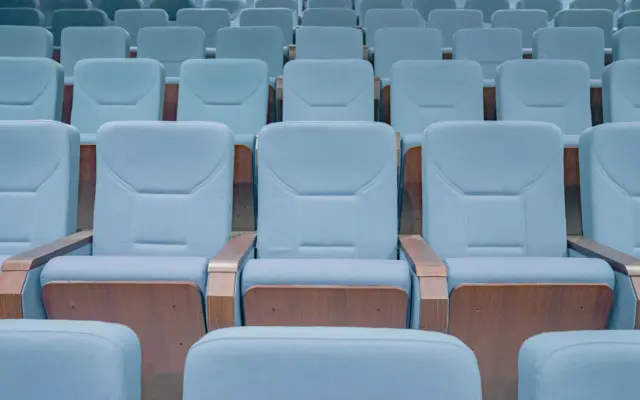 Rânduri succesive de scaune de culoare bleu, cu brațe din lemn, dintr-o sală de cinema sau amfiteatru