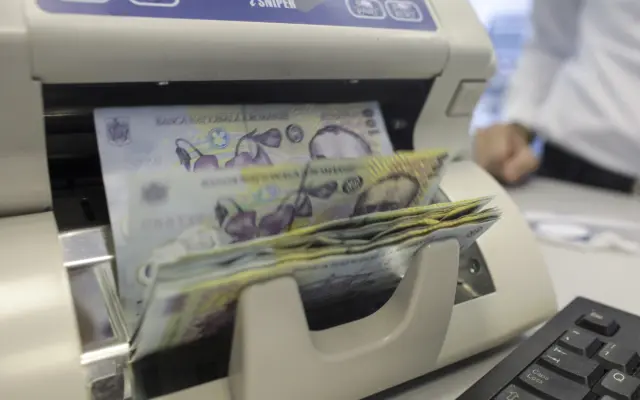O mașină de numărat bani (aparat iSNIPER) prin care trec teancuri de bancnote de 100 de lei românești (RON), ilustrând fluxurile financiare și banii publici